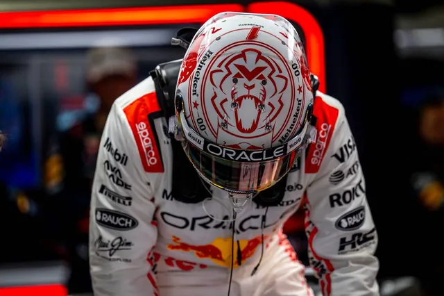 Verstappen in the garage in white race overalls, helmet on, head lowered.
